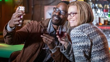 Man and woman taking a selfie photograph with a mobile phone while both holding a glass of red wine.
