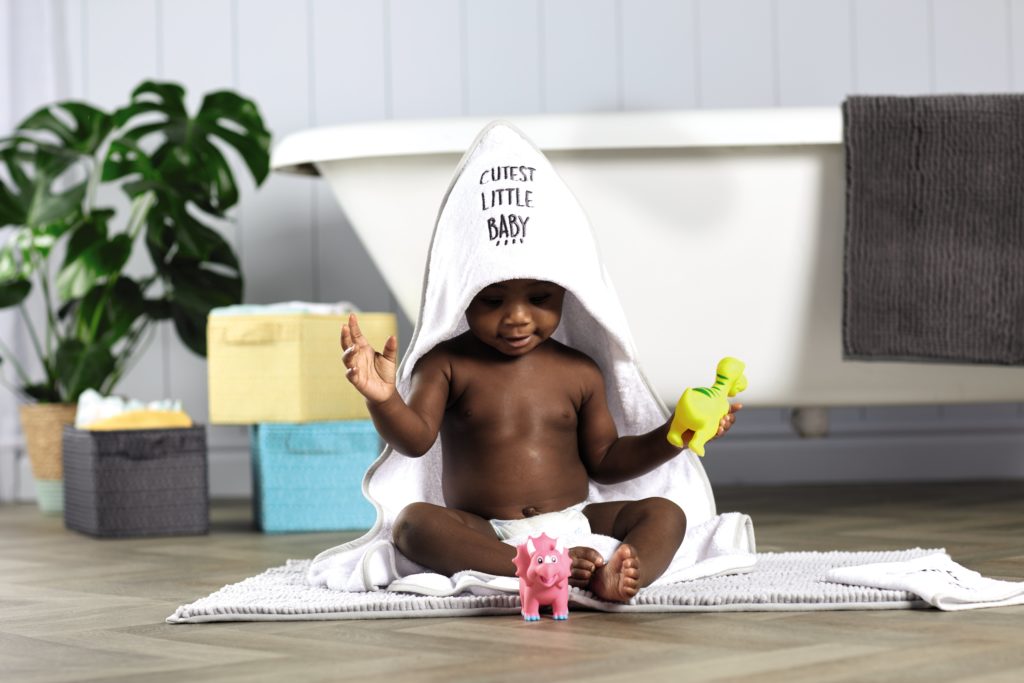 A baby enjoying bath time wrapped in a white hooded towel and playing with brightly coloured bath time toys 