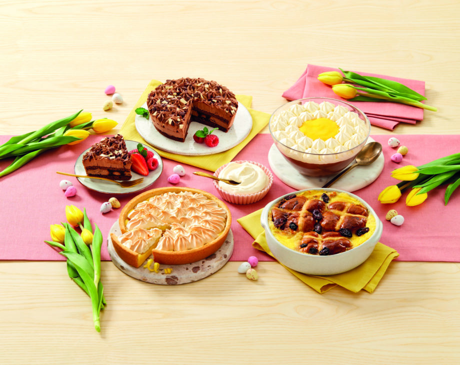 A range of Aldi's Easter desserts arranged on a table, laid on a pink tablecloth and surrounded by yellow tulips.
