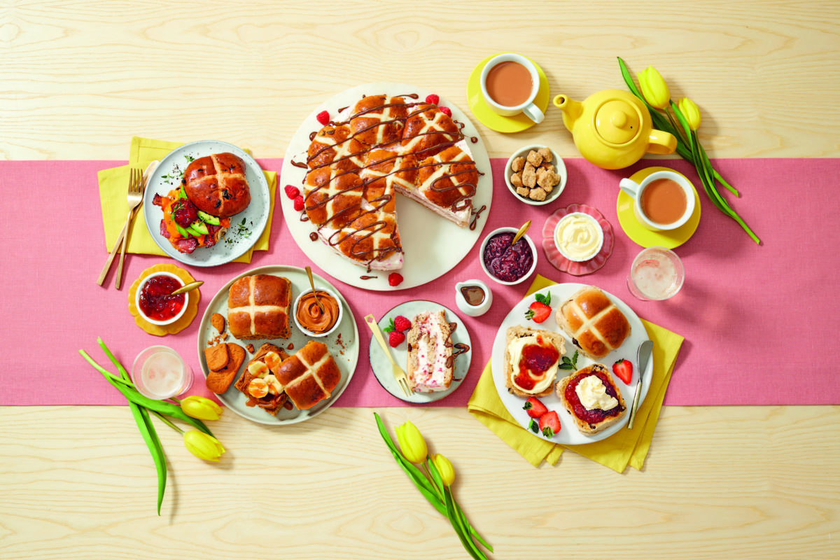 Aldi's range of Hot Cross Buns displayed on plates. Sitting atop a pink tablecloth and surrounded by yellow tulips and yellow tea pots.