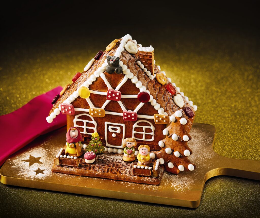 DIY gingerbread house. Fully assembled with white icing decoration for the windows and doors. Slanted roof with sweets for decoration. On a gold board.