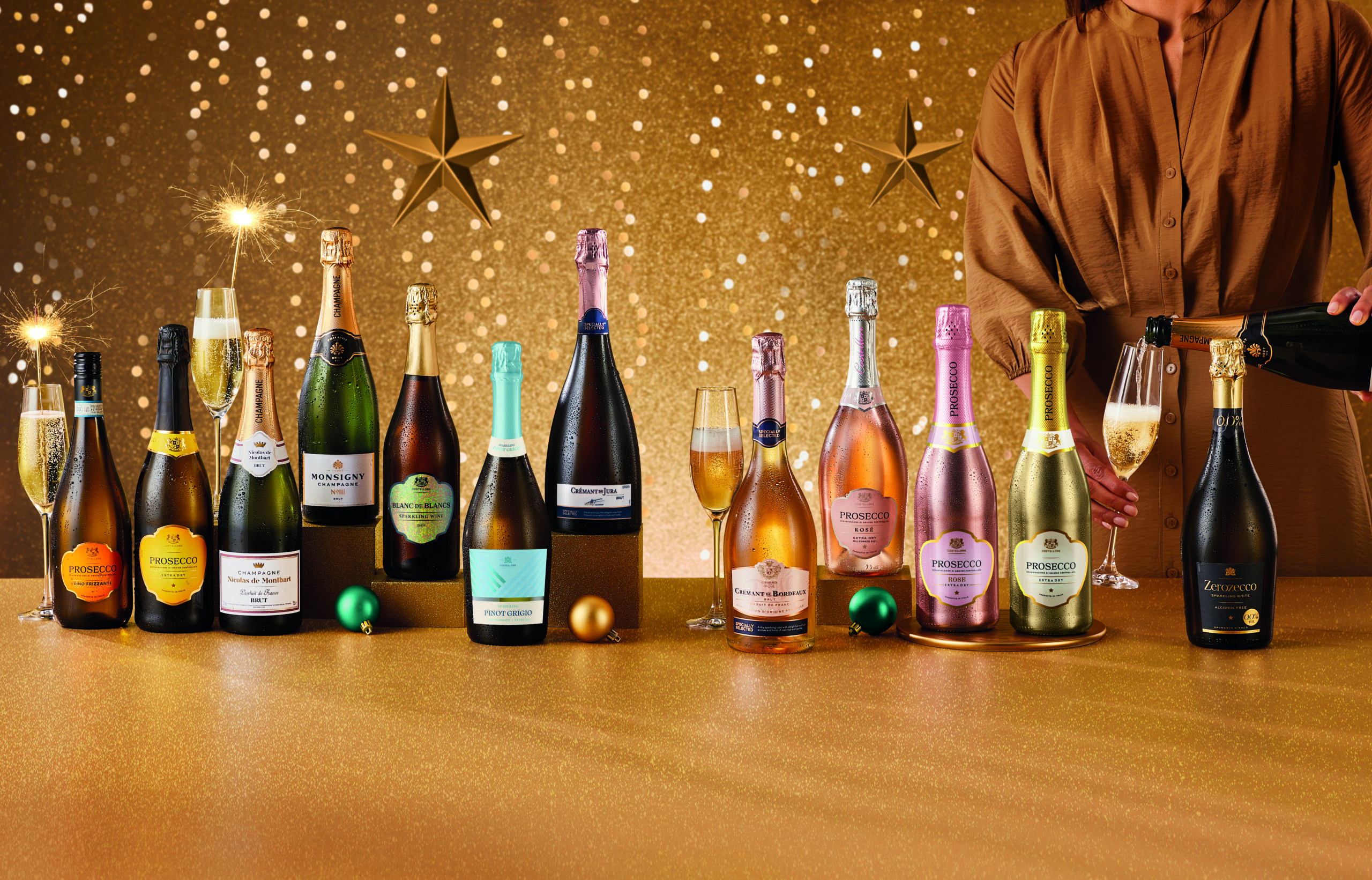 A gold background with fairylights and star decorations, with an array of bottles of fizz with sparklers sticking out