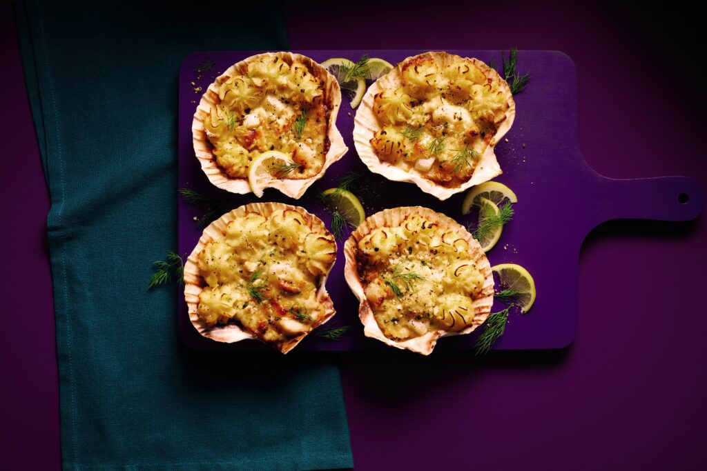 four scallop gratins displayed in shell halves on a purple board