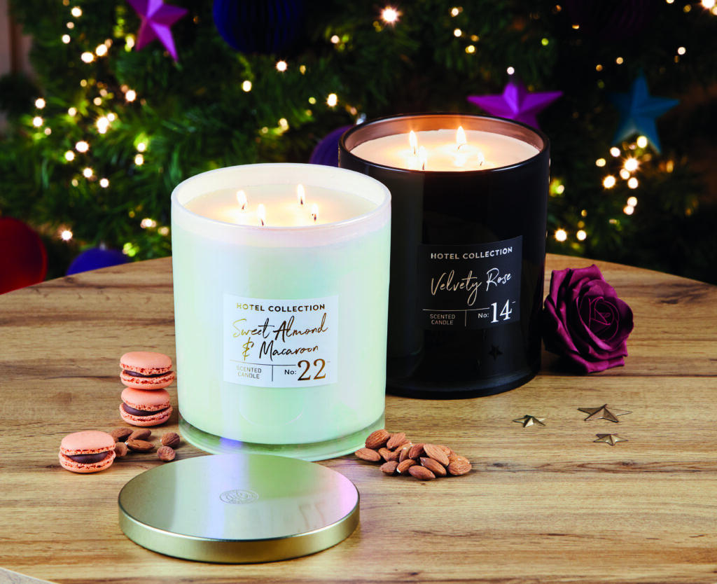 Two candles, one white and one black are light on a wooden table in front of a Christmas tree. Next to the candles are macarons and flowers.